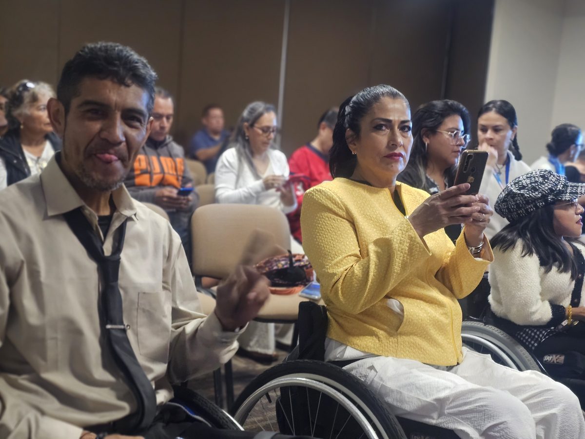 Asistentes a un evento en un auditorio; en primer plano, un hombre sonríe a la cámara.