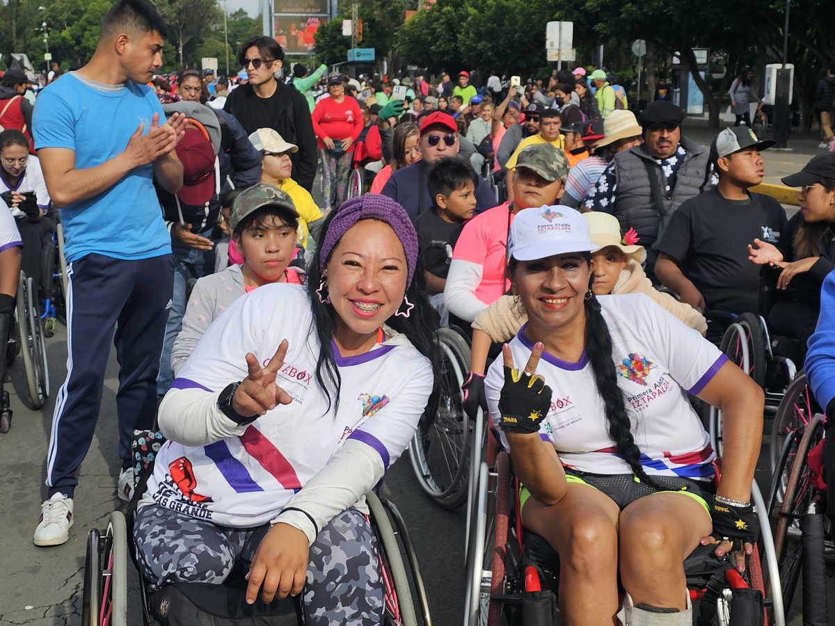 Dos mujeres en silla de ruedas, con playeras de un evento, saludan a la cámara entre una multitud al aire libre.